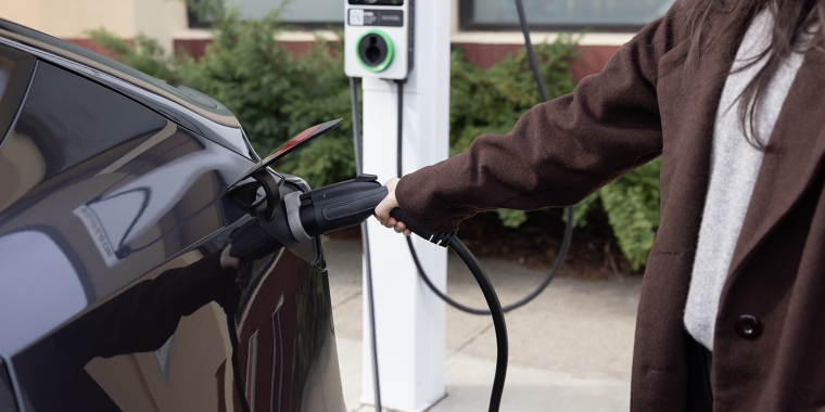 A person in a brown coat is charging an electric car at a BAT electric vehicle charging station. The charging cable is plugged into the car, and greenery is visible in the background.